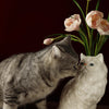 A gray tabby cat gently nuzzles a white ceramic cat sculpture vase holding pink tulips.