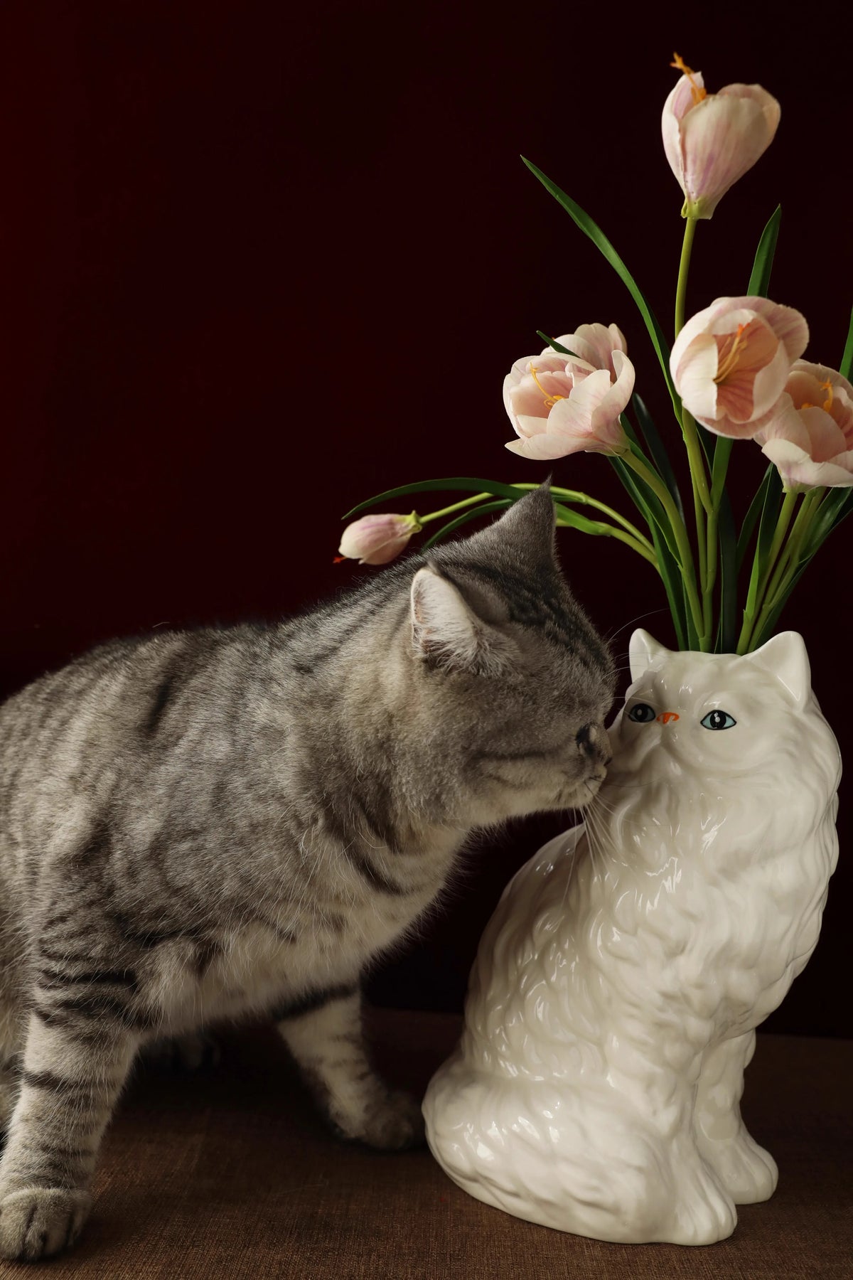A gray tabby cat gently nuzzles a white ceramic cat sculpture vase holding pink tulips.