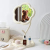 A white floral decorative mirror with a rounded frame reflects a teddy bear and is placed on a light-colored surface.