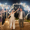 A bride and groom are celebrating outdoors with lighted tinsel wedding wands creating sparkling light effects.
