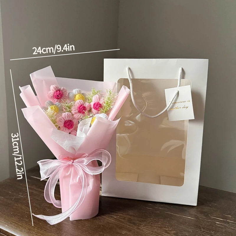 A pink handmade crochet flower bouquet with yellow accents is displayed in a white gift bag.
