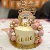 A first birthday cake decorated with pink ball and gold accents, featuring a rainbow topper and the name Julia.