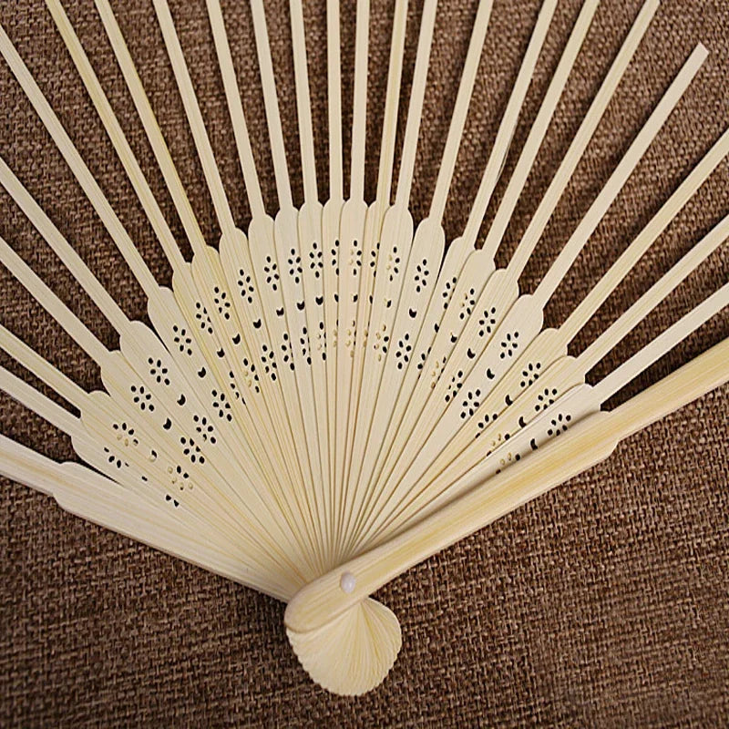 Ventilateur à main en bambou jaune clair pliable avec perforations délicates, exposé sur un tissu marron.