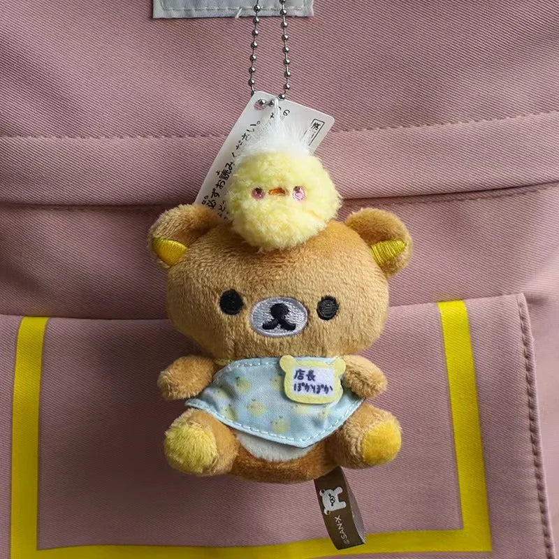 A fluffy brown bear plush keychain with a yellow nose and a light blue bib hanging from a string against a pink fabric backgr
