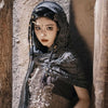 A female model wearing a dark hooded cloak with distressed fabric and a Viking-style brooch, looking directly at the camera.
