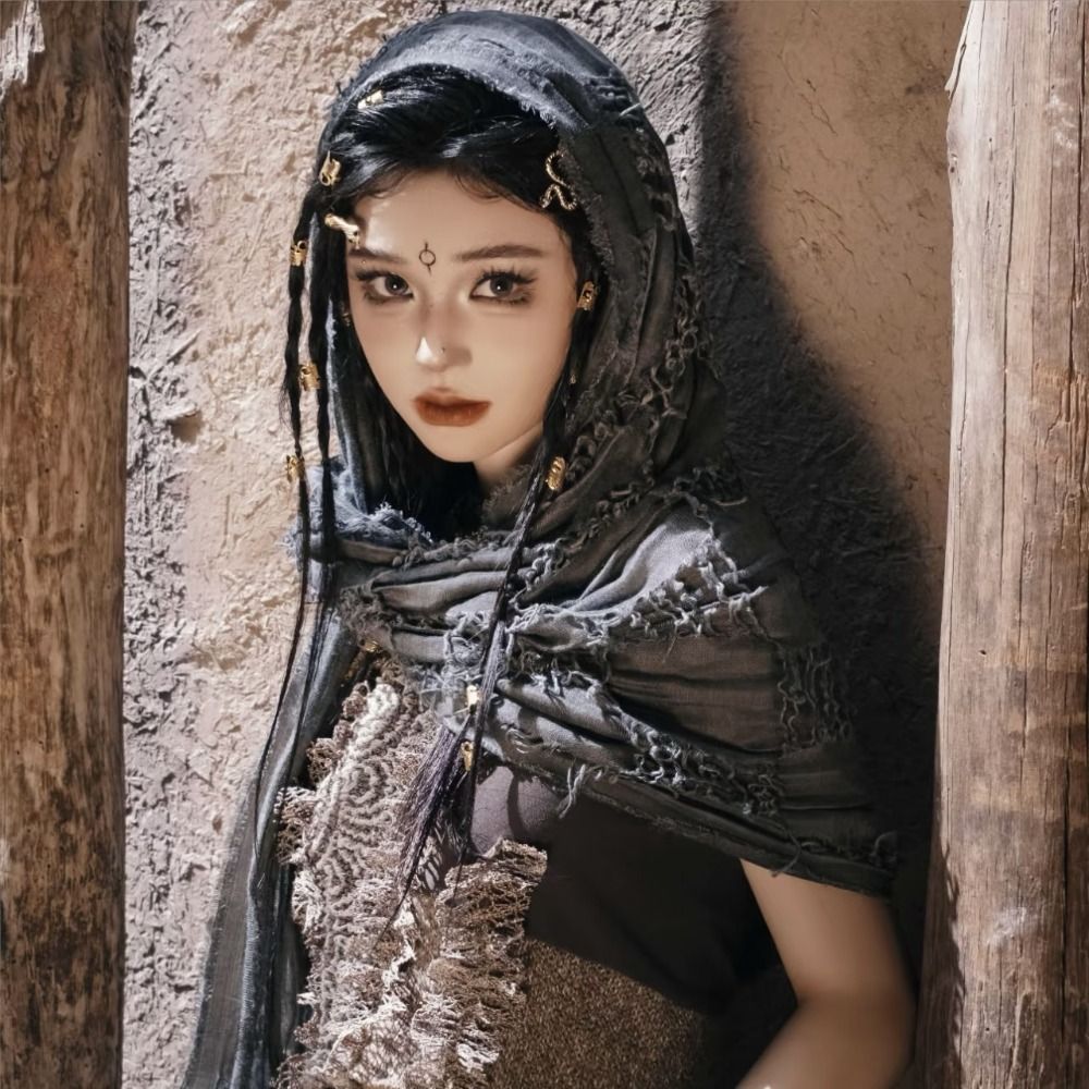 A female model wearing a dark hooded cloak with distressed fabric and a Viking-style brooch, looking directly at the camera.