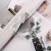 A clear acrylic sheet with a vintage clock design and eucalyptus leaves is placed on a white surface next to a camera.