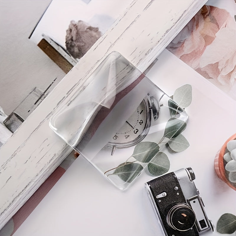 A clear acrylic sheet with a vintage clock design and eucalyptus leaves is placed on a white surface next to a camera.